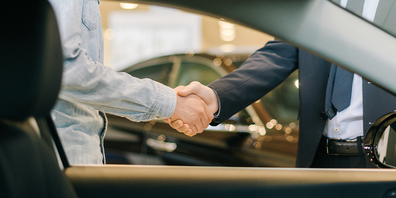 Close-up of a car salesperson shaking hands with a customer framed in a vehicle's window
